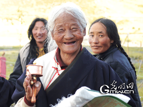 A Tibetan senior in traditional Tibetan costume swings her prayer wheel with a smile, photo from Gmw.cn.