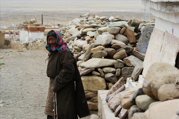 An elder Tibetan woman stands by a Mani stone pile. [Photo/www.dili360.com]